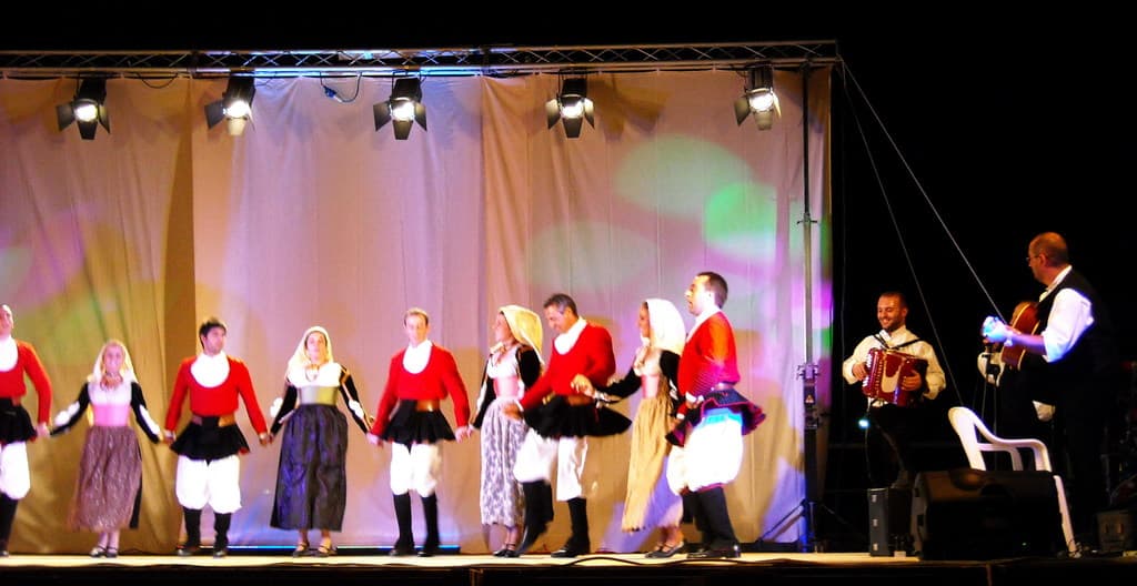 Leistende auf der Bühne mit einem Vorhang im Hintergrund, Lampen oben, Musiker rechts und ein weißer Stuhl in der rechten unteren Ecke.