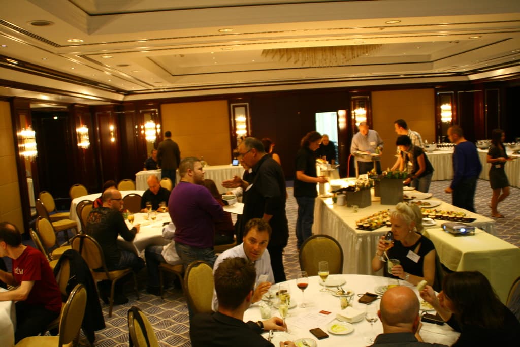 Ein Restaurant-Innenraum mit Tischen und Stühlen, Gruppen von Menschen beim Essen und einem zentralen Tisch mit verschiedenen Speisen, einige Menschen stehen und essen.