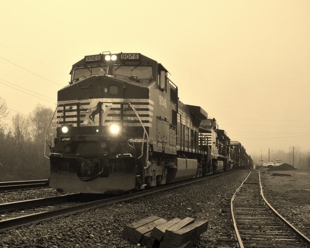 Ein Zug auf Schienen mit Autos, Strommasten, Bäumen und Himmel im Hintergrund.