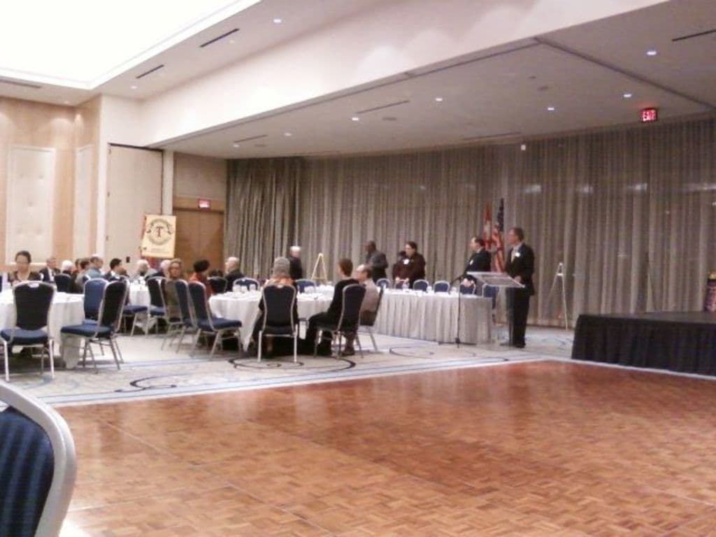 Eine Gruppe von Menschen, die auf Stühlen sitzen, einige stehen, Fahnen auf Stangen, Lichter, Schilder und Vorhänge im Hintergrund.