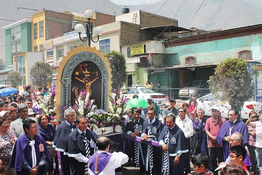 Menschen auf einer StraÃe vor der Stadt, mit einem Kruzifix und BlumenstrÃ¤uÃen in der Mitte, fahrende Autos dahinter und GebÃ¤ude, BÃ¤ume und StraÃenlaternen im Hintergrund.