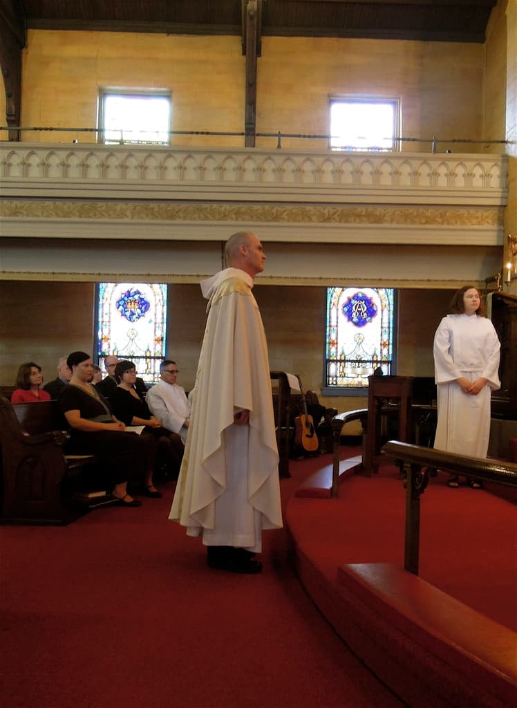 Eine Frau und ein Mann in weißen Kleidern stehen in einer Kirche, mit einem roten Teppich, Bänken, Musikinstrumenten und Fenstern im Hintergrund.