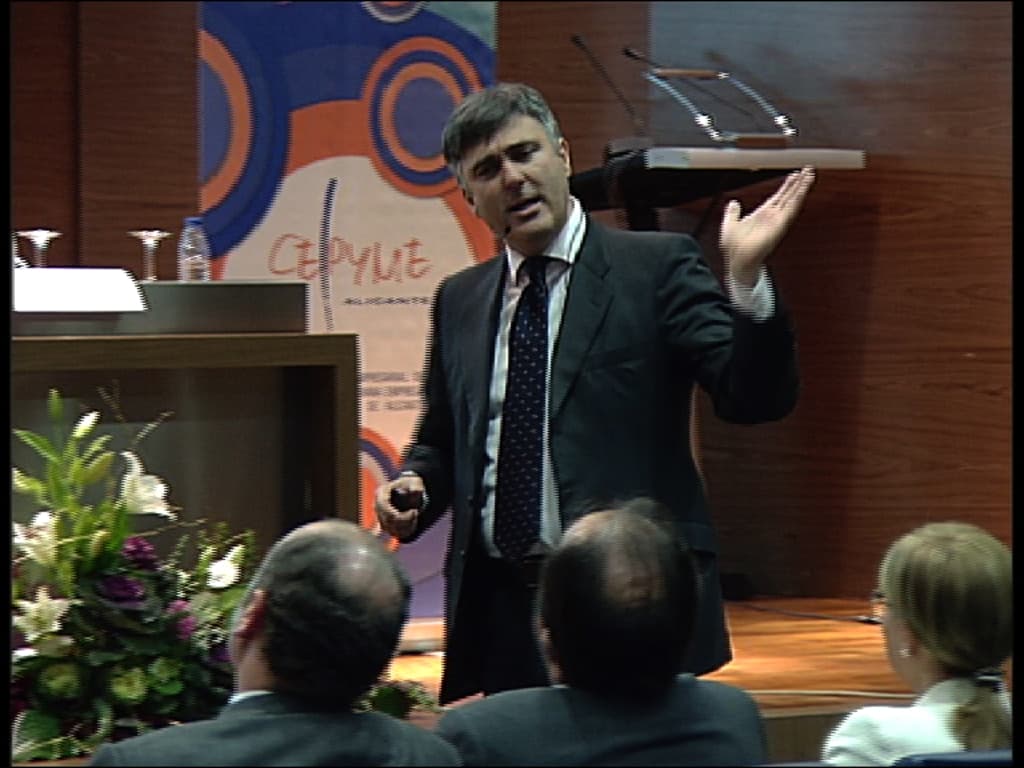 Drei Personen sitzen in der ersten Reihe eines Konferenzsaals, ein Mann in einem schwarzen Blazer steht und spricht in ein Mikrofon, ein Blumenarrangement und ein Tisch mit einem Buch und einer Wasserflasche in der Nähe, ein Podium mit Mikrofonen dahinter und eine farbenfrohe Tafel mit Text im Hintergrund.