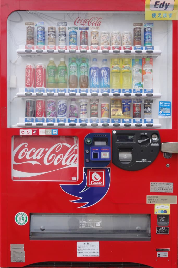 Ein Coca-Cola-Automat mit Flaschen, die oben hinter einer Glasscheibe angeordnet sind und Stangen, die unten am Automaten befestigt sind.