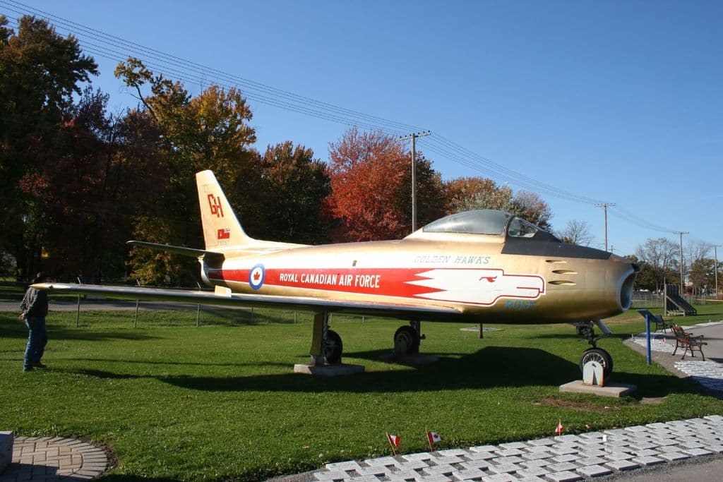 Ein Flugzeug steht auf dem Boden mit einem Mann in einer Mütze, Jacke, Jeans und Schuhen auf der linken Seite, einer kleinen Flagge unten, Bänken auf der rechten Seite und einem Hintergrund aus Bäumen, Strommasten und Drähten unter einem Himmel.
