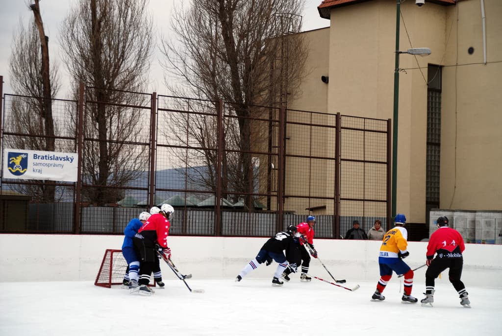Personen beim Eishockey auf einem Eisring mit Gebäuden, Bäumen, einer Straßenlaterne, einem Namensschild und Zäunen im Hintergrund unter einem Himmel.
