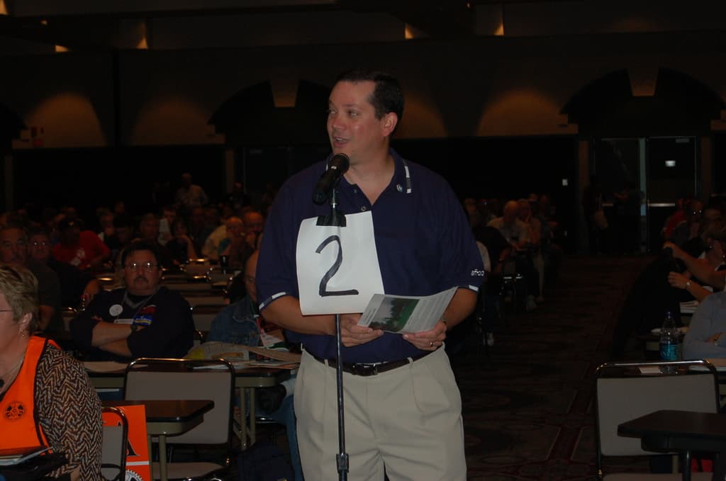 Innenansicht eines Stadions: Ein Mann in einem blauen T-Shirt spricht in ein Mikrofon auf einem Ständer, hält ein Blatt Papier in der Hand und hat Gruppen von Menschen zu seiner Linken und Rechten auf Stühlen sitzend, eine Wasserflasche und ein Buch auf einem Tisch sowie eine Person, die vor einer Tür steht, im Hintergrund.