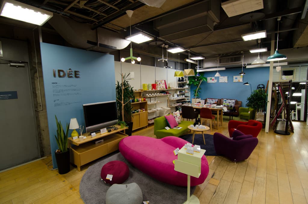 Eine Ladeneinrichtung mit Haushaltsgeräten und Möbeln, darunter ein Fernseher auf einem Tisch, eine rosafarbene Couch, ein Sofa, zwei Stühle um einen kleinen Hocker, ein Esstisch mit Stühlen und verschiedene Gegenstände auf Regalen, alles beleuchtet von Deckenlampen.