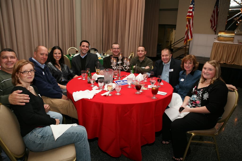 Eine Gruppe strahlender Menschen sitzt an einem Tisch mit Gläsern, Papieren, Weinflaschen und -gläsern, einer Flagge auf der rechten Seite und Vorhängen an der Wand.