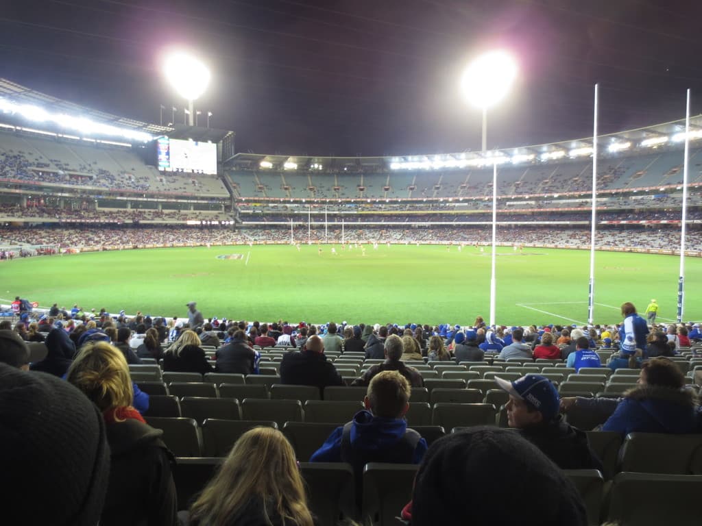 Ein Stadion mit Rasen, vielen auf Stühlen sitzenden Menschen, Leuchten im Hintergrund, Pfosten auf der rechten Seite und einem Bildschirm im Hintergrund.