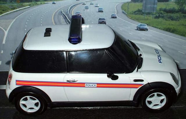 Ein weißer Polizeiwagen steht im Vordergrund mit vielen Autos, die sich auf der Straße dahinter bewegen, umgeben von Gras und Bäumen auf beiden Seiten.