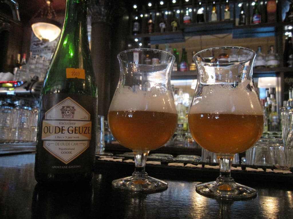 Eine Flasche mit Logo und zwei Weingläser auf einem Tisch, umgeben von vielen Gläsern, Flaschen und Gestellen.