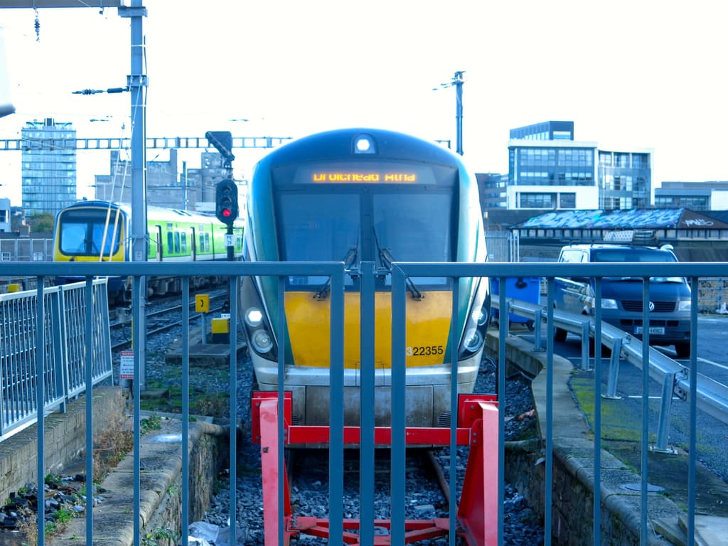 Zwei Züge auf den Schienen mit einem Zaun im Vordergrund, einige Pfosten auf dem Land einschließlich einem mit einer Signalleuchte, ein Auto auf der Straße rechts, Gebäude im Hintergrund und der Himmel oben im Bild.