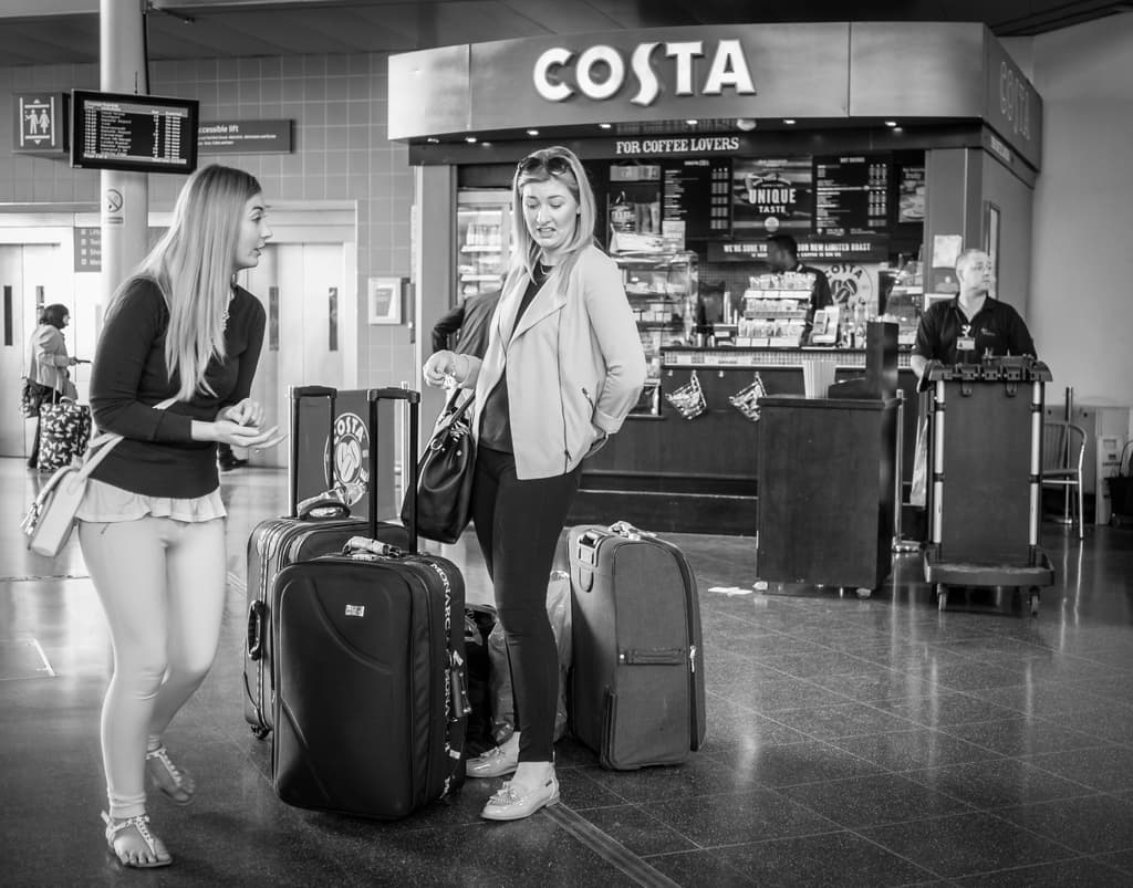 Zwei Frauen mit Gepäck vor einem Café in einem scheinbar Flughafen stehend.