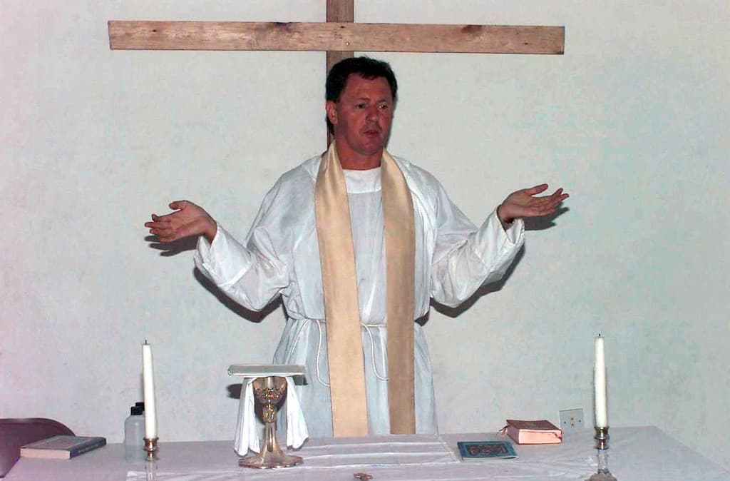 Ein Priester steht in der Mitte des Bildes mit einem Tisch vor ihm, auf dem Kerzen und Bücher liegen.