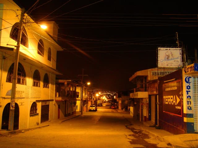 Eine Straßenansicht mit Gebäuden und Strommasten auf beiden Seiten, einigen parkenden Fahrzeugen vor den Gebäuden und einer Straße in der Mitte.