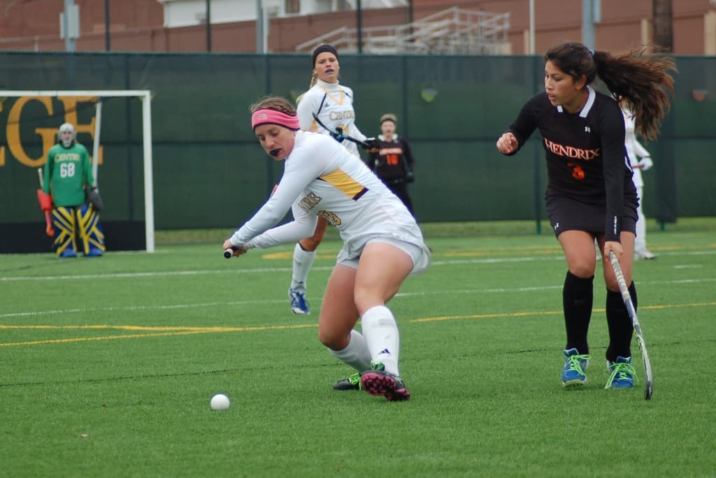 Eine Gruppe von Menschen spielt Hockey auf Gras, wobei eine Frau in der Mitte versucht, einen weißen Ball mit ihrem Stock zu treffen, während andere ihre Stöcke in der Nähe halten, und verschiedene Ausrüstungsgegenstände und Strukturen im Hintergrund sichtbar sind.