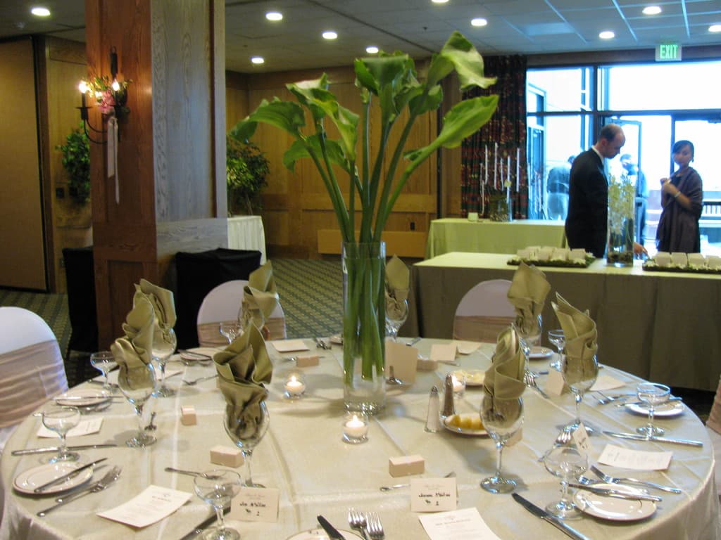 Ein Tisch mit einer weißen Decke, Tellern, Besteck und einem Glas, umgeben von Stühlen, mit einem grünen Tisch dahinter; ein Mann in einem schwarzen Blazer und ein Mädchen stehen in der Nähe einer Ausgangstür, mit zwei Männern dahinter.