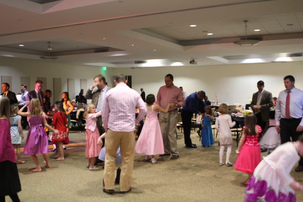 Eine Gruppe von Kindern und Erwachsenen, die um Tische mit Gegenständen darauf, Stühlen und von der Decke hängenden Lampen stehen.