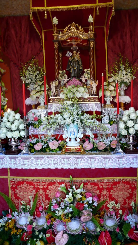 Idole auf einem Tisch mit Blumenvasen und Kerzenständern, die Kerzen halten, mit Blumen und Blättern unten und Vorhängen im Hintergrund.