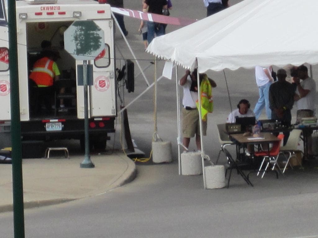 Ein Krankenwagen steht auf der linken Seite einer Straße, während auf der rechten Seite ein Zelt viele Menschen beherbergt, mit einem Tisch und einem Stuhl in der Nähe.