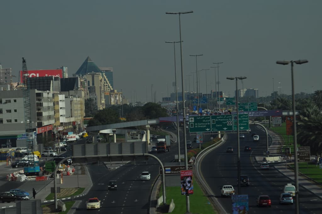 Eine Luftaufnahme einer Stadt mit Gebäuden, Türmen, Kraftfahrzeugen, Werbetafeln, Schildern, Straßen, Bäumen, Straßenlaternen, Menschen, Verkehrskegeln und Informationsanzeigen unter einem klaren Himmel.
