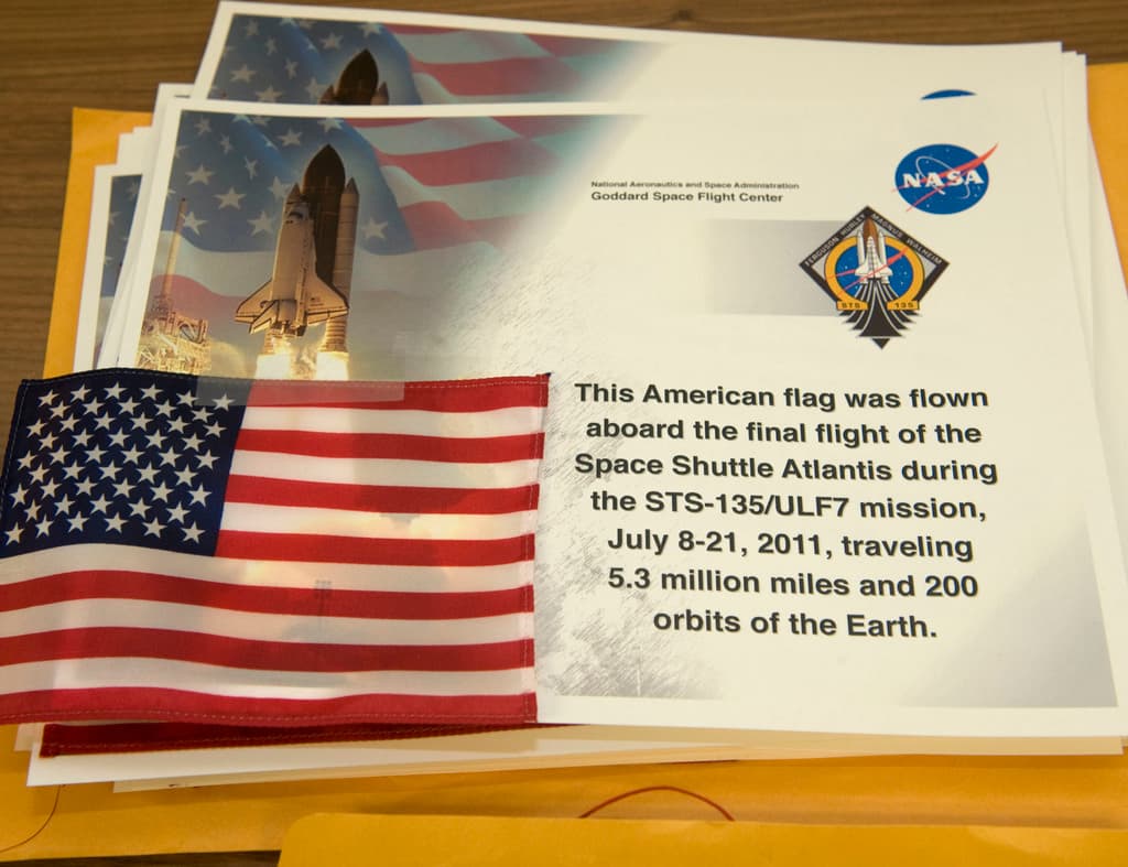 Plakate mit Text, einer Flagge, einer Rakete und Logos sind in der Mitte zu sehen, mit einem Tisch unten und einem Boden oben im Bild.