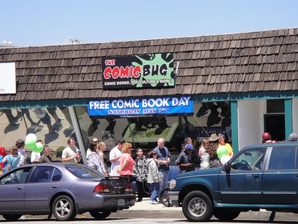 Gruppen von Menschen vor einem Gebäude mit Glastüren und einem Dach, zwei Autos auf der Straße, grüne Luftballons, ein Banner und eine Namensschild auf dem Dach, unter einem klaren Himmel.