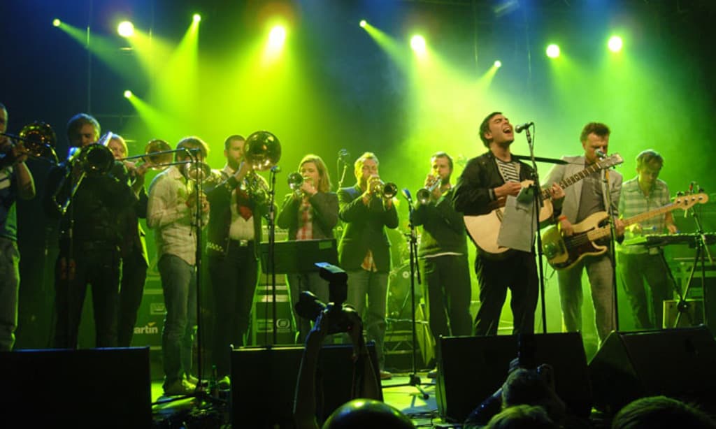 Eine Gruppe von Musikern performs auf der Bühne mit Mikrofonen, Lichtern und einer Kamera, während das Publikum zusieht.