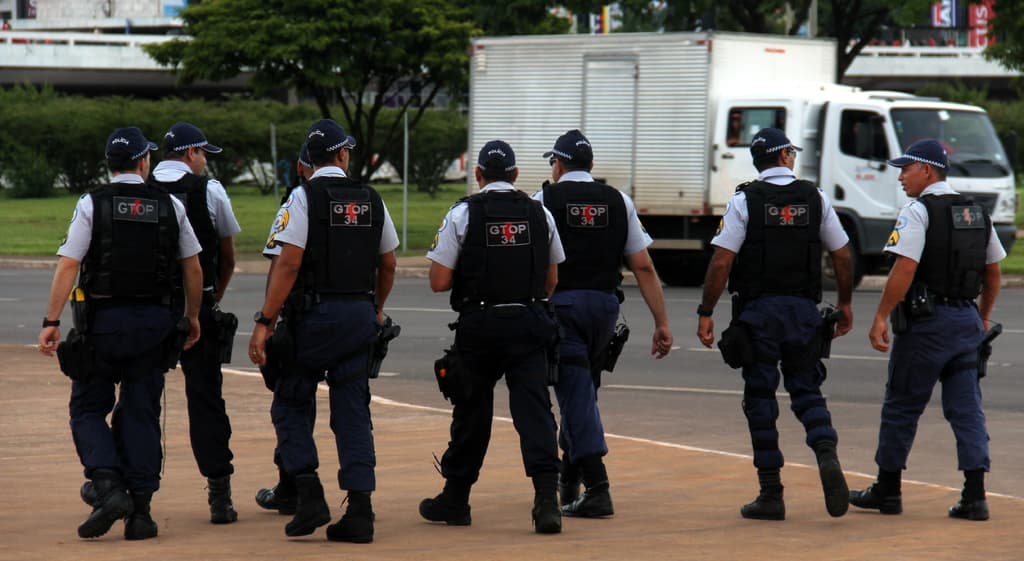 Eine Gruppe von Polizisten in Uniform, einschließlich Mützen, Hemden, kugelsicheren Westen, Uhren, Hosen und Schuhen, geht auf einer Straße. Rechts daneben steht ein Lastwagen in der Nähe eines Zauns, mit Gebäuden, Bäumen, Pflanzen, Pfählen und Bannern im Hintergrund und einer Brücke oben rechts.