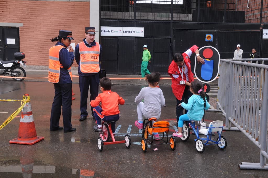 Kinder auf Fahrrädern auf einer Straße mit Verkehrshütchen, Menschen, Geländern auf der rechten Seite, einem Fahrrad auf der linken Seite, einer Wand mit Türen im Hintergrund und Schildern.