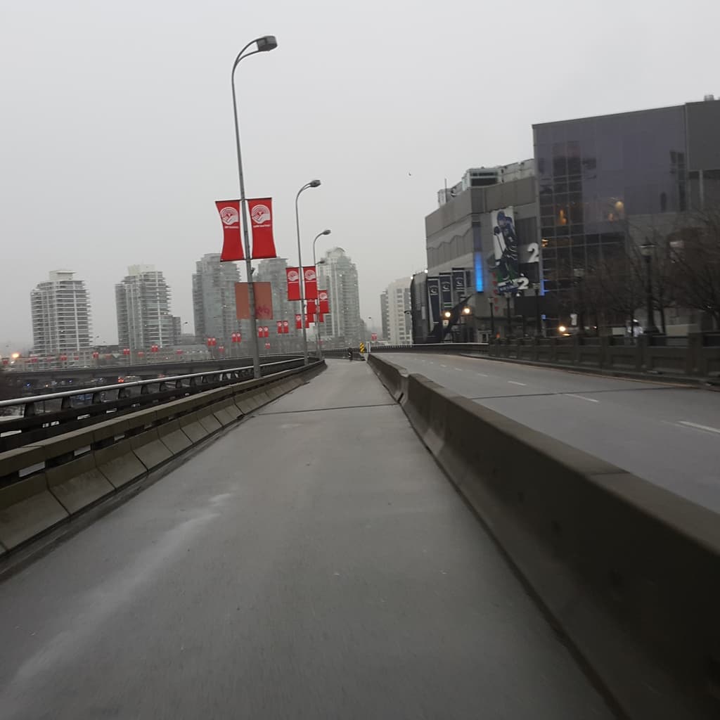 Eine Ansicht von Straßen mit Straßenlaternen daneben, Gebäuden im Hintergrund, einem Himmel darüber und einem Geländer links.