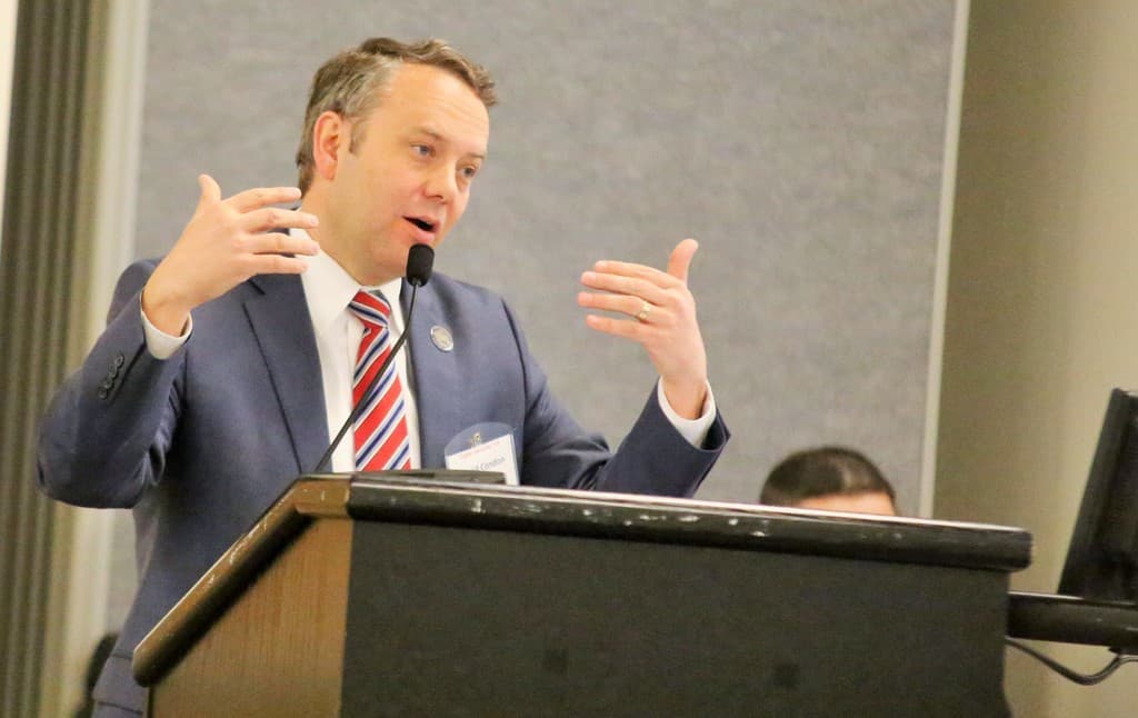 Ein Mann steht vor einer Wand mit verschiedenen Gegenständen auf einem Podium mit einem Mikrofon.