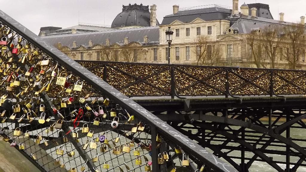 Eine Trestelbrücke mit zahlreichen Schlössern daran, umgeben von Bäumen und Gebäuden im Hintergrund.