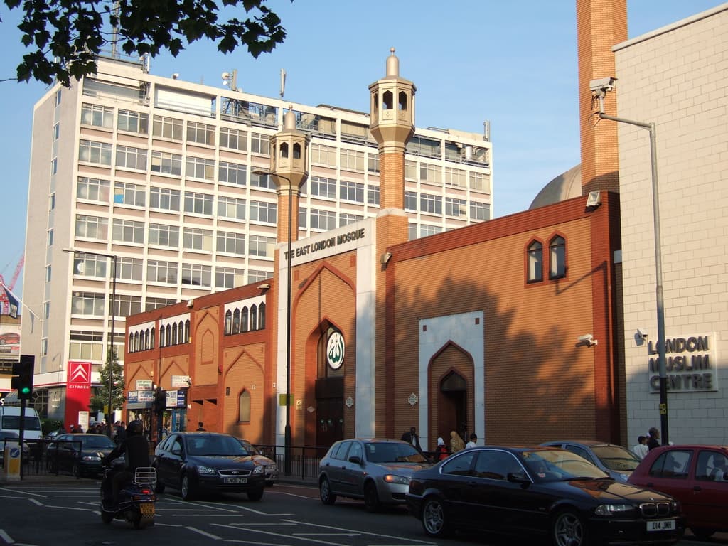 Eine Moschee mit einem Namensschild an der Wand, Straßenlaternen, eine Ampel an einem Mast, Fahrzeuge auf der Straße, Gruppen von Menschen, die stehen, ein Baum und eine an einem Mast befestigte Überwachungskamera.