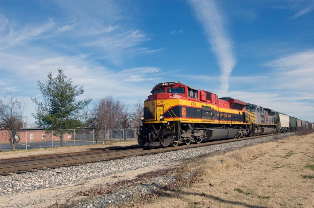 Ein Zug auf der rechten Seite des Bildes, mit einer Bahnstrecke in der Mitte, Boden unten, Zaun links, Bäume im Hintergrund und Himmel oben.