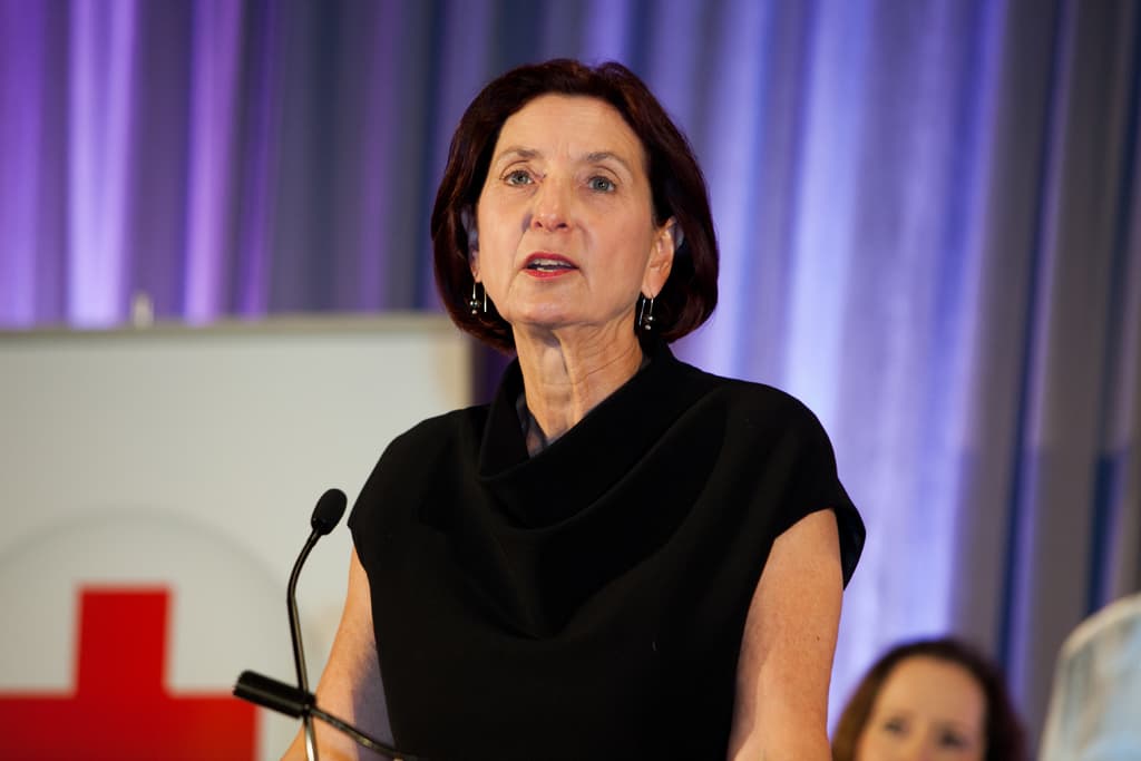 Eine Frau in einem schwarzen T-Shirt steht an einem Mikrofon und hält eine Rede, mit einer sitzenden Frau dahinter, einer weißen Tafel mit einem Kreuzsymbol und einem violetten Vorhang im Hintergrund.