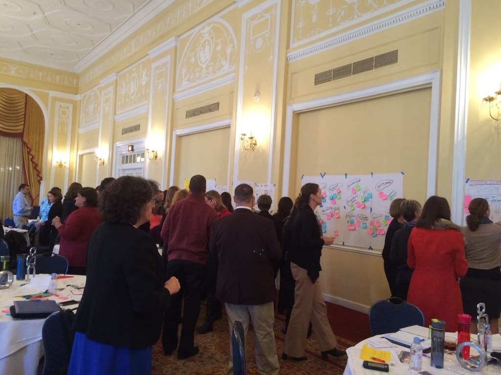 Eine Gruppe von Menschen betrachtet Plakate und Charts, die an einer Wand in einem Raum zu sein scheinen, der wie ein Kunstmuseum aussieht.