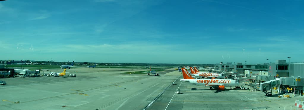 Eine Luftaufnahme eines Flughafens mit Flugzeugen, Gebäuden, Polen, Fahrzeugen, Bäumen, einer Straße unten und einem Zaun im Vordergrund.