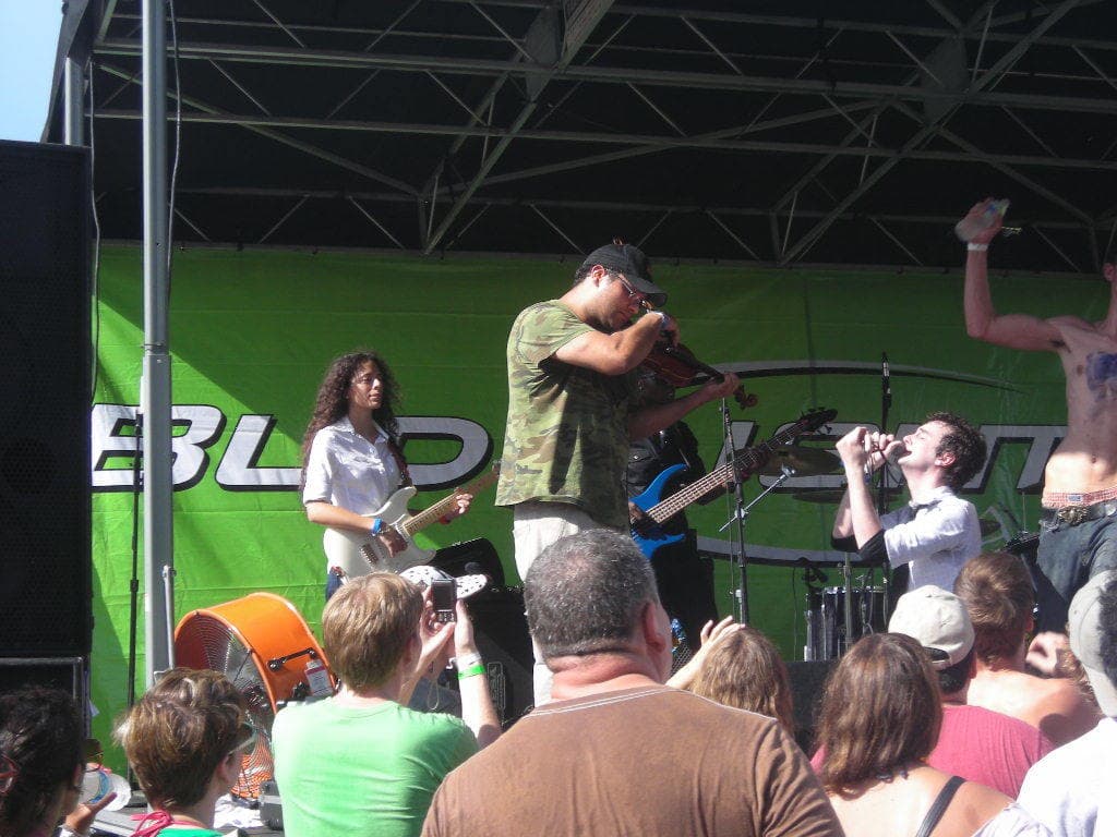 Eine Gruppe von Musikern, die verschiedene Instrumente auf einer Bühne spielen, während das Publikum davor steht und die Vorstellung genießt, und ein grüner Banner im Hintergrund.