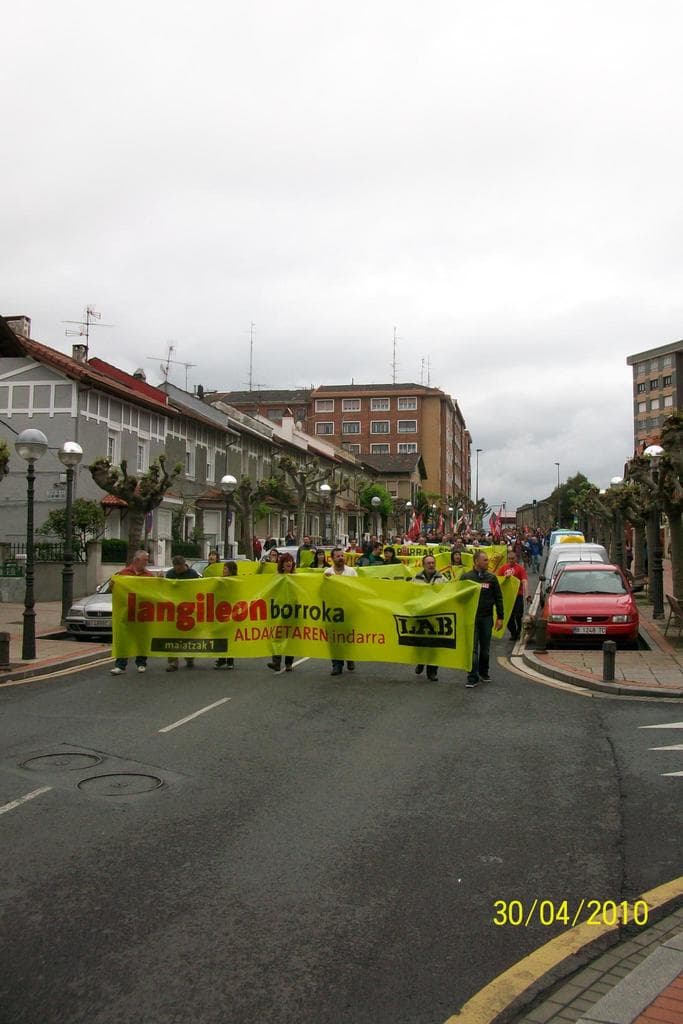 Eine Gruppe von Menschen hält Schilder und marschiert auf einer Stadtstraße, mit Fahrzeugen, Polen und Bäumen auf den Gehwegen, Gebäuden im Hintergrund und einer bewölkten Himmel.