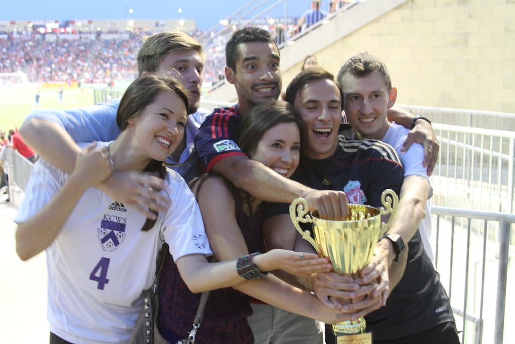 Sechs Menschen, darunter zwei Frauen und vier Männer, halten einen Pokal und lächeln, während sie für ein Foto vor einem Eisengeländer posieren, mit einer Wand und einem Geländer oben rechts und Menschen und Lichtern im Hintergrund, möglicherweise in einem Stadion.