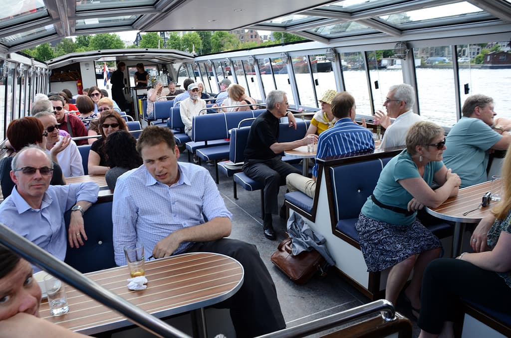 Ein Boot-Innenraum mit Sitzreihen, die von Menschen besetzt sind, Tischen mit Gläsern und Schüsseln davor, einer Tasche auf dem Boden, Wasser im Hintergrund, Gebäuden auf der rechten Seite und kleinen Pflanzen über dem Boot.