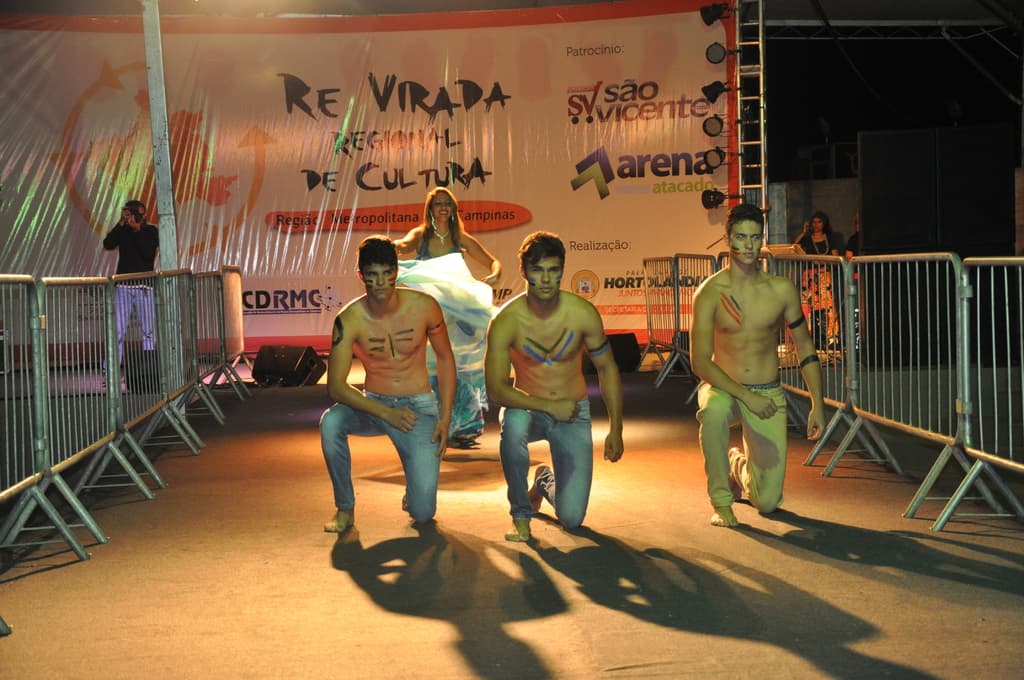 Drei Jungen ohne Hemden knien auf dem Boden während eines kulturellen Ereignisses, mit Grills auf beiden Seiten und einem großen Banner im Hintergrund, während eine Person mit einer Kamera auf der linken Seite ein Foto macht.