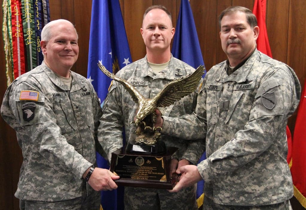 Drei Männer in Uniformen halten Auszeichnungen, mit Fahnen dahinter und bunten Gegenständen mit Text in der oberen linken Ecke.