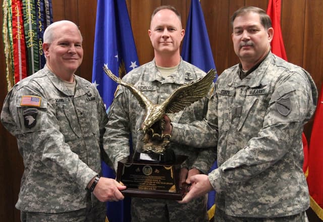 Drei Männer in Uniformen halten Auszeichnungen, mit Fahnen dahinter und bunten Gegenständen mit Text in der oberen linken Ecke.