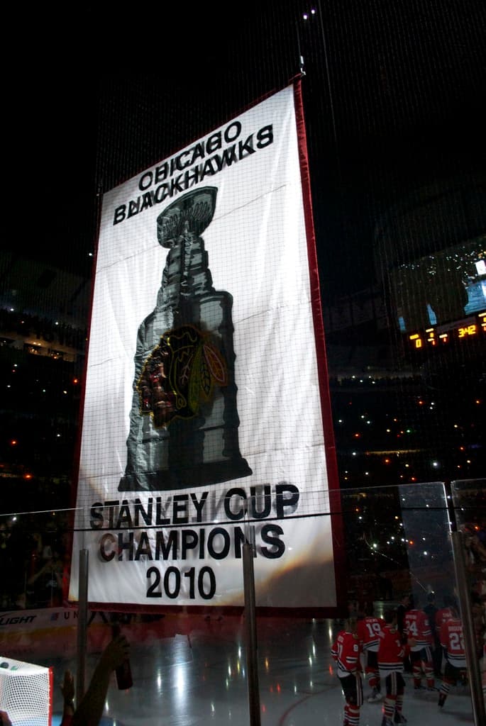 Eine Nachtstimmung im Stadion mit einem groÃen Banner in der Mitte, Zuschauern im Hintergrund, einem Glas auf dem Boden und einigen Spielern auf dem Feld, beleuchtet von Stadionlüstern.
