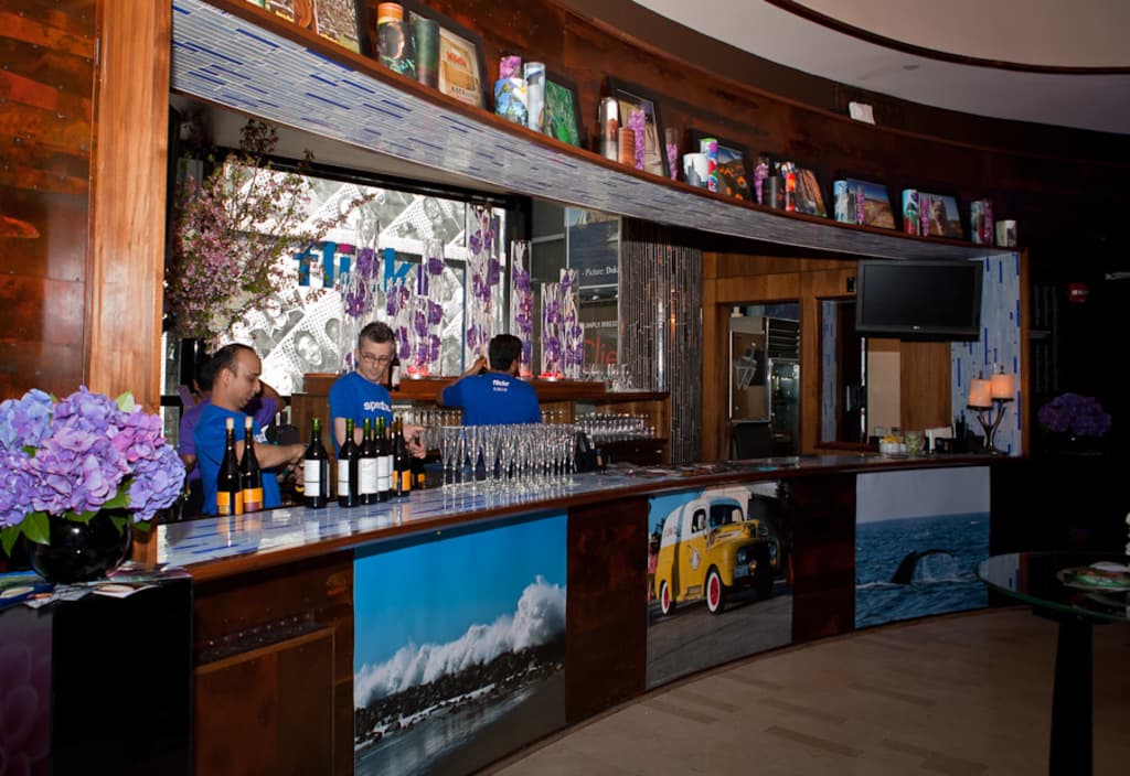 Ein Restaurantinnenraum mit drei Personen, die hinter einem Tisch mit Gläsern, Flaschen, Aufklebern, Postern und einer Zimmerpflanze arbeiten, mit einem Fernseher und Lampen sowie Gegenständen auf einem Regal in der Nähe.