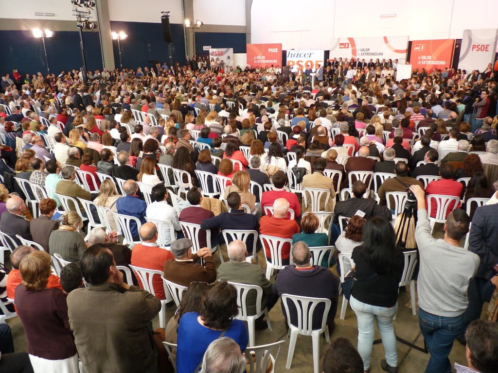 Ein volles Stadion mit vielen sitzenden und stehenden Menschen, eine Bühne mit zwei Rednern an einem Podium und mehreren Fahnen und Lichtern in der Nähe.