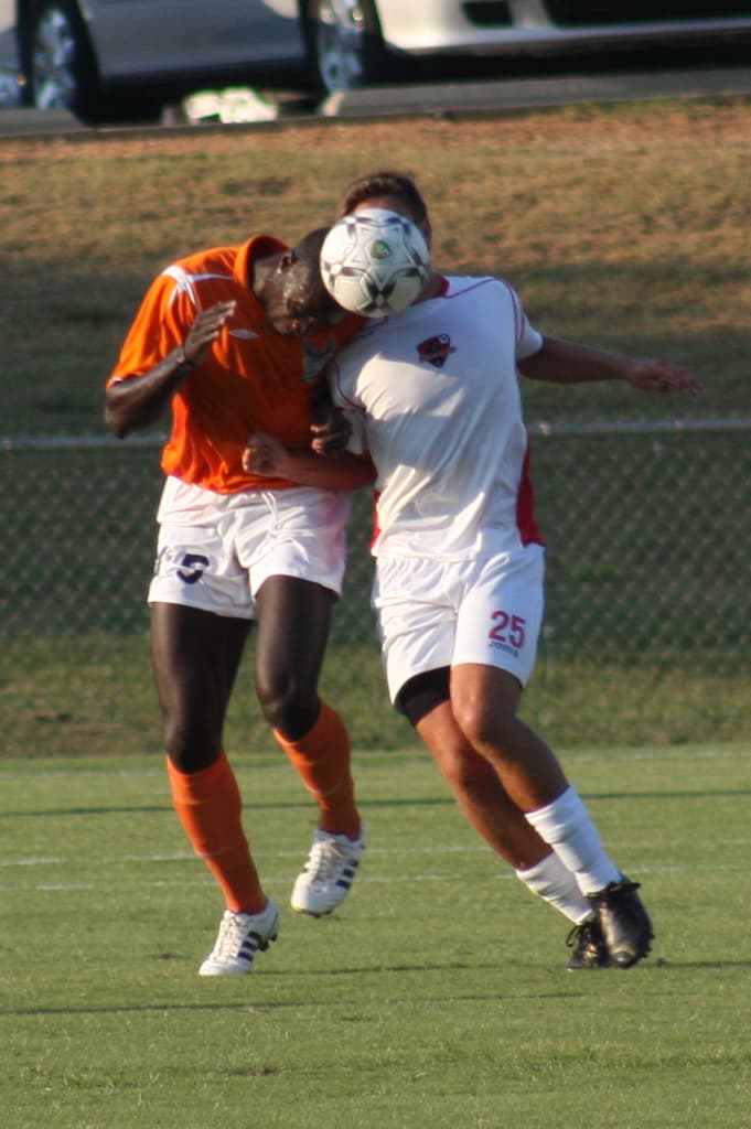 Zwei Spieler auf einem umz√§unten Platz, einer in einem orangen T-Shirt, der den Ball mit dem Kopf sch√§gt, mit Autos und Gras im Hintergrund.